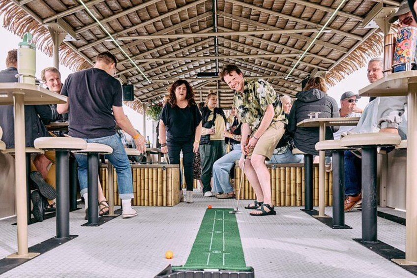 A group of people playing a game with a ball on a green mat under a thatched roof structure. 3