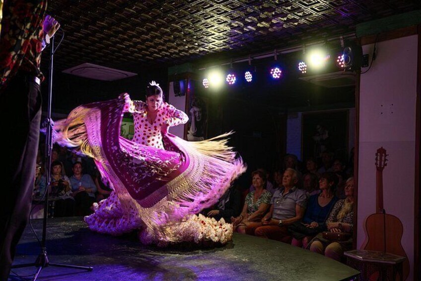 Un artista con un vestido llamativo baila en el escenario mientras el público observa en una sala con poca luz. 3