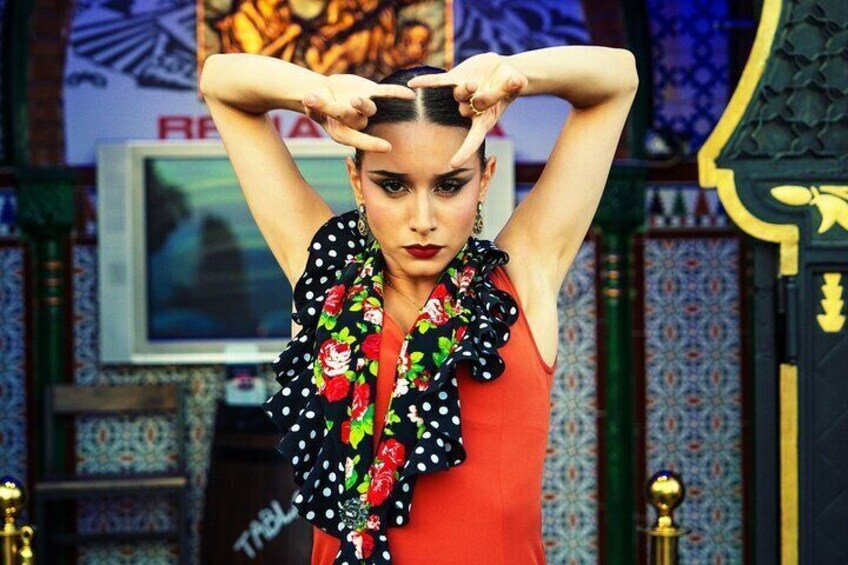 Una persona con vestido rojo y una bufanda blanco y negro a lunares, de pie frente a una pared con diseño, un televisor y objetos decorativos. 1