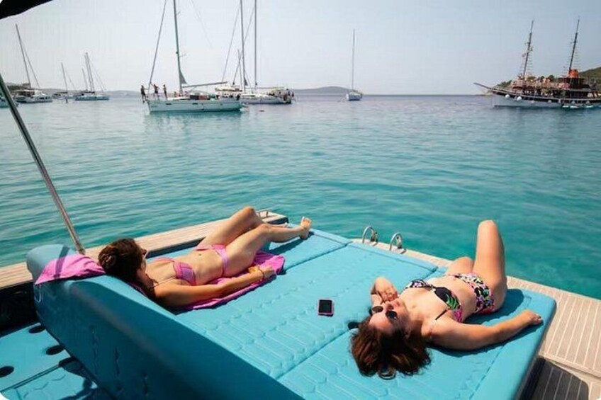 Two people sunbathing on a boat deck with a clear blue sea and sailboats in the background. 4