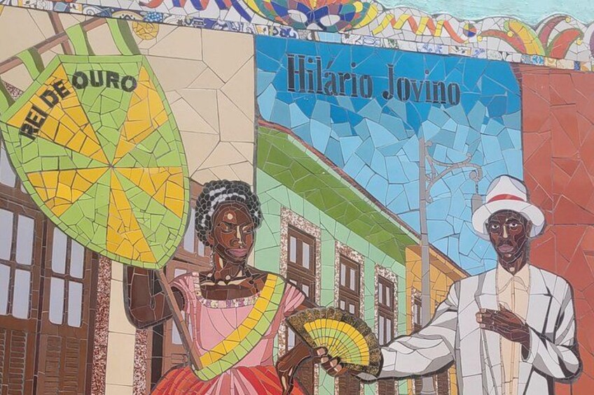 Un murale a mosaico raffigura figure che reggono ventagli e un edificio con un cartello che riporta la scritta 'Hilario Jovino'. 5