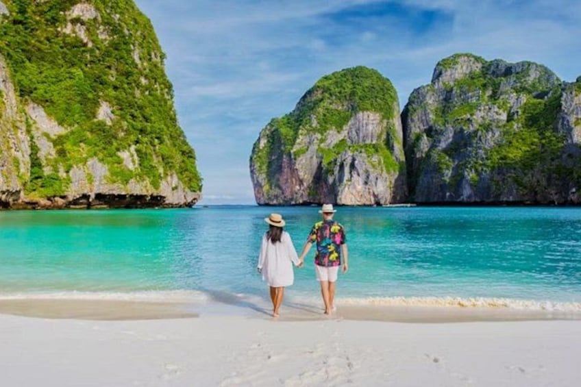 Una playa con dos personas caminando sobre la arena, un mar azul cristalino y grandes formaciones rocosas verdes al fondo. 3