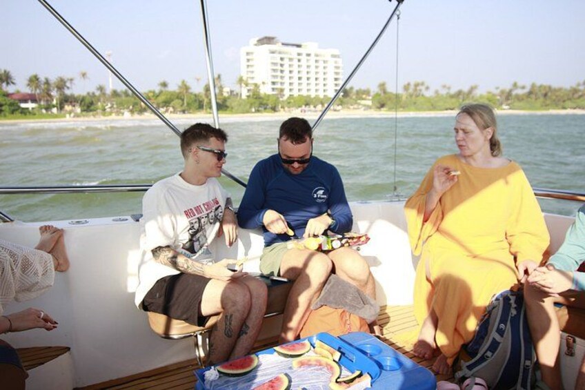 A boat with people sitting and relaxing, a beach with palm trees, and a building in the background. 1