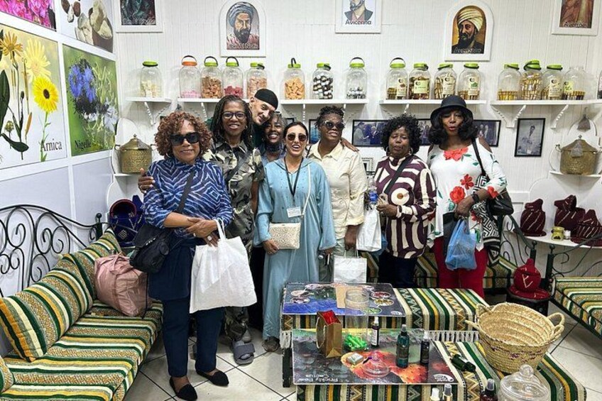 A group of people in a room with patterned sofas, a table with various items, and shelves with jars and bags. 4