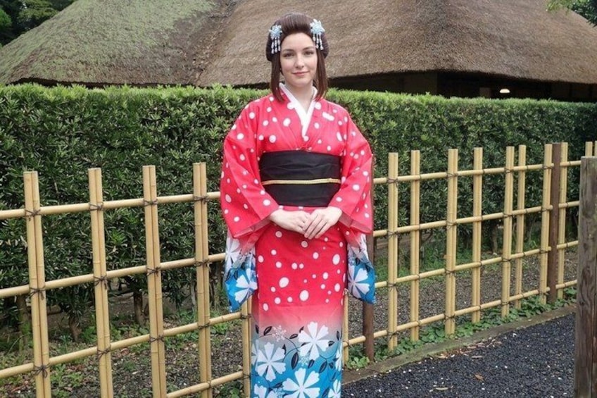 A person in traditional Japanese attire standing in front of a bamboo fence with a thatched-roof structure in the background. 4