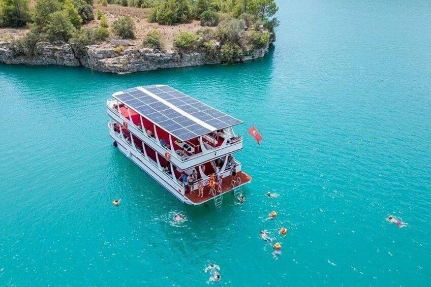 A double-deck boat with solar panels on a turquoise body of water surrounded by greenery. 1