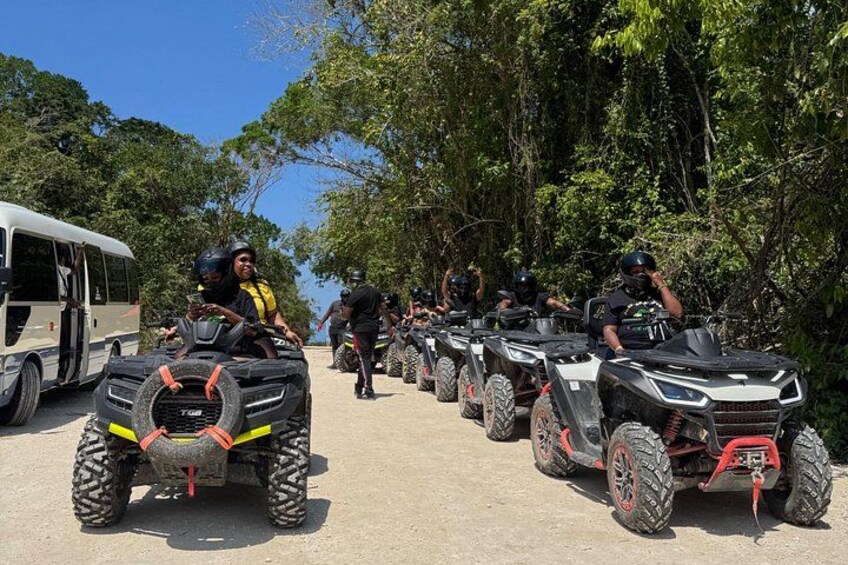 A group of people on ATVs lined up on a dirt path with trees and a bus in the background. 2
