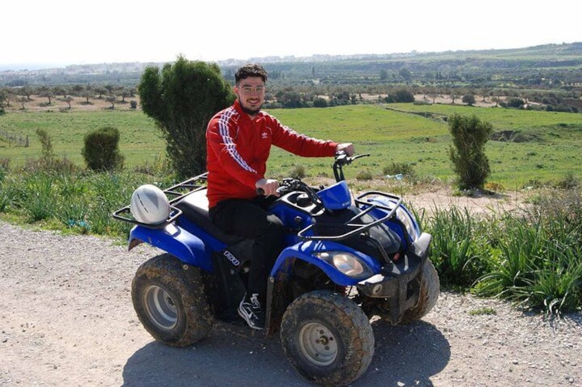 A person riding a blue ATV on a dirt path with green fields and trees in the background. 2