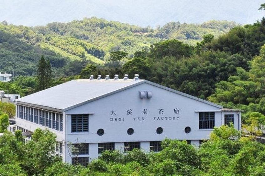 Ein weißes Gebäude mit der Aufschrift 'DAXI TEA FACTORY' auf der Fassade, umgeben von üppiger Vegetation und Hügeln im Hintergrund. 4
