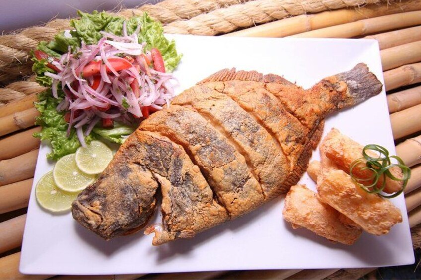 Ein ganzer frittierter Fisch auf einem weißen Teller, dazu Zitronenspalten, ein Beilagensalat und gebratene Kochbananen. 3