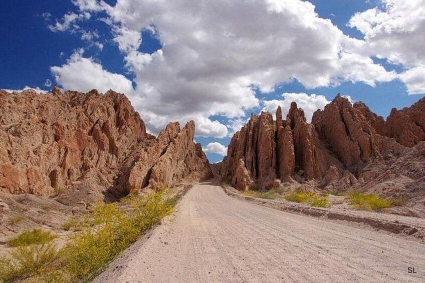 A dirt road flanked by rocky cliffs under a partly cloudy sky. 1