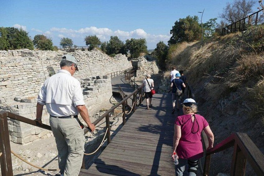 A wooden boardwalk with people walking, stone ruins, and a grassy hillside. 4