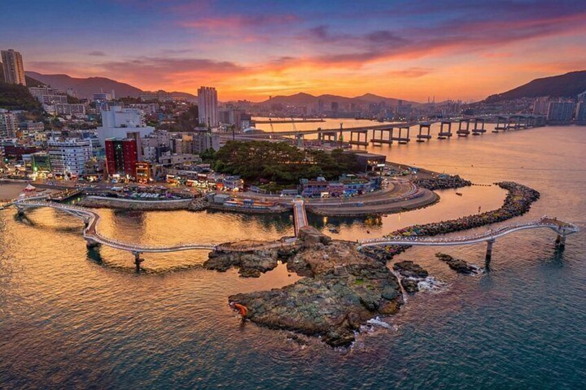 Un paisaje urbano costero al atardecer con un puente destacado, una península con edificios y un litoral rocoso. 5