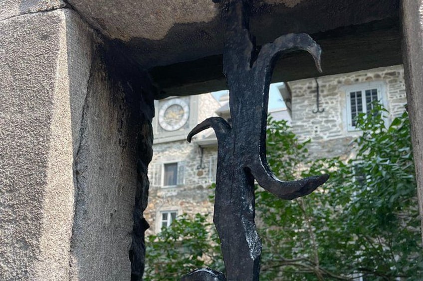 A wrought iron hook attached to a stone structure, with a stone building and greenery in the background. 5
