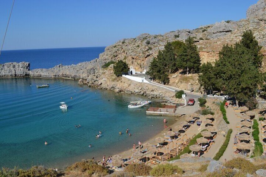 A coastal area with clear blue waters, rocky cliffs, and a beach with umbrellas and people. 5