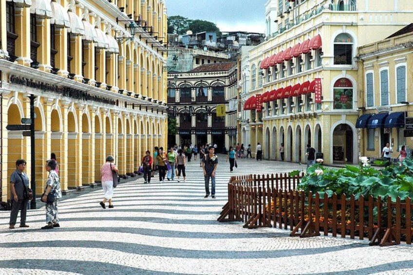 A pedestrian street lined with yellow buildings, a central open space, and a wooden fence with greenery. 2