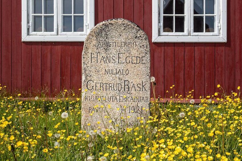 A gravestone with inscriptions in front of a red wooden building. 3