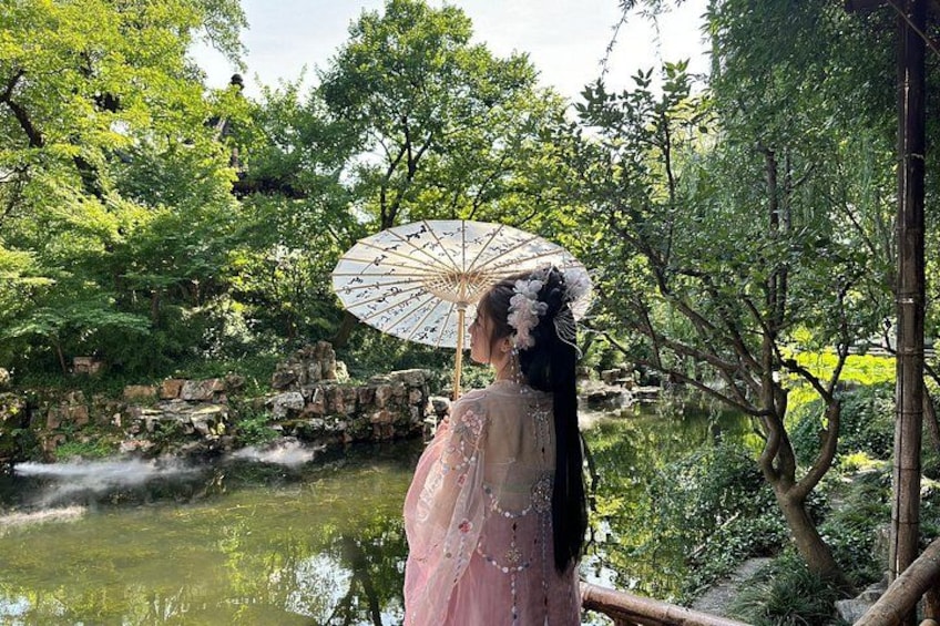 A person in traditional attire holding an umbrella, standing by a pond with trees and rocks in the background. 5