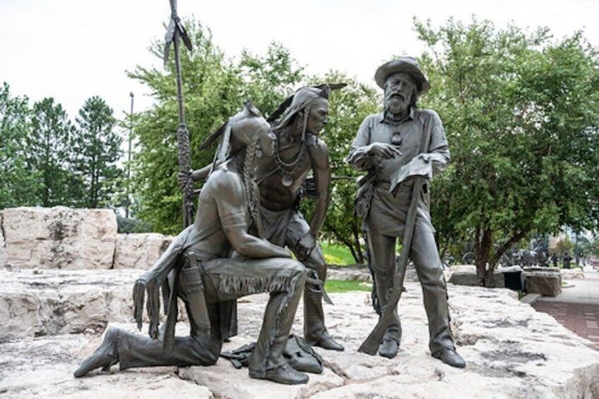 A statue of three figures in historical attire, one seated and two standing, in an outdoor setting with trees and stone structures in the background. 3