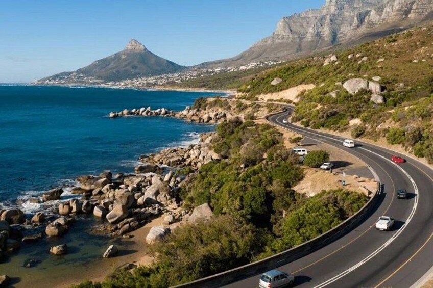A coastal road with a mountain in the background, surrounded by greenery and rocky terrain. 1