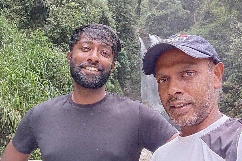 Two individuals standing in a natural setting with dense greenery and a waterfall in the background. 4