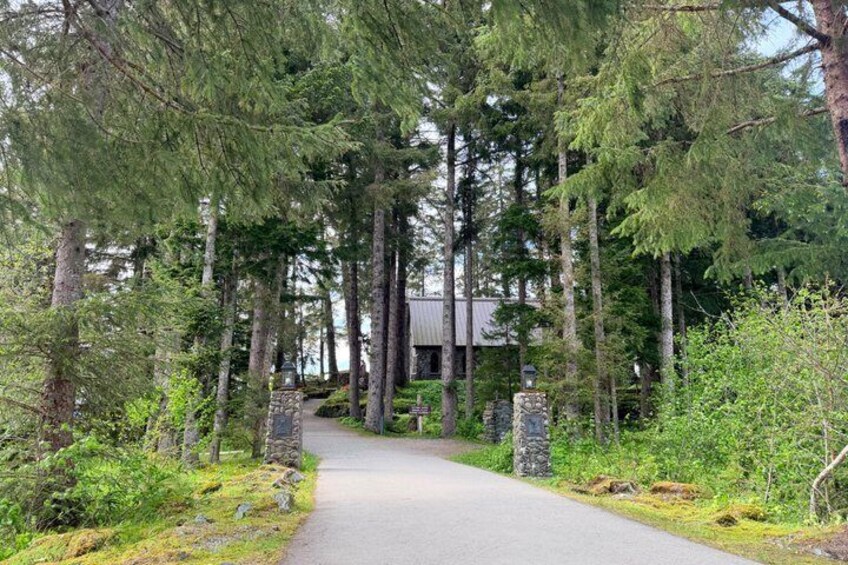 A paved path through a forest with tall trees and a building in the background. 5