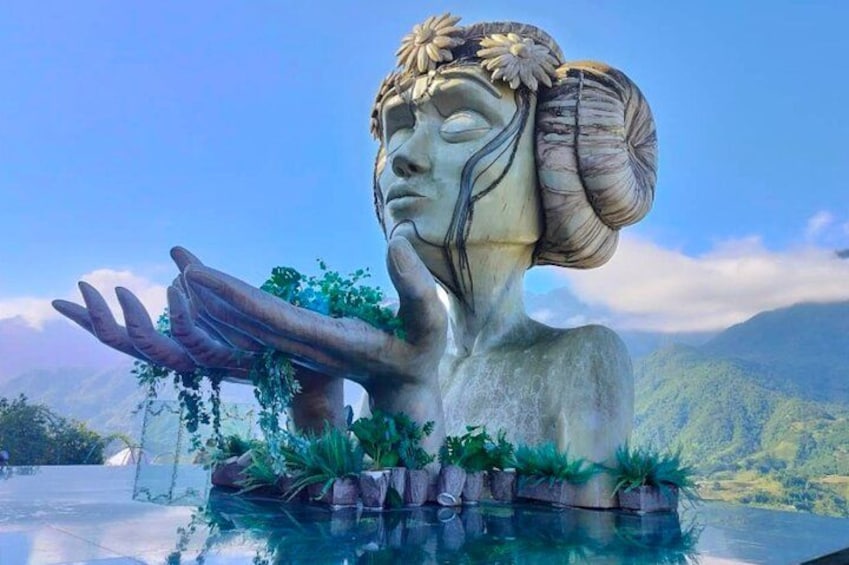 A statue with a hand extended, surrounded by greenery and water, set against a mountainous backdrop. 3