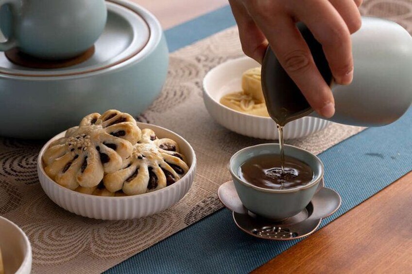 Una mano vertiendo té en una taza, un cuenco de pasteles y una tetera sobre una mesa. 4