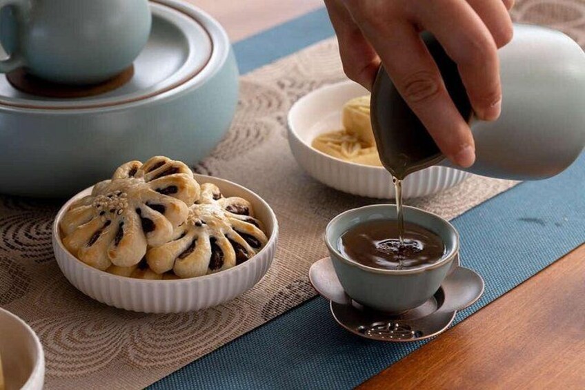 A hand pouring tea into a cup, a bowl of food, and a teapot on a table. 4