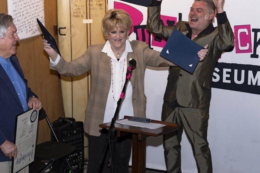 A person standing at a podium with a microphone, holding a folder, and another person raising their hand. 2