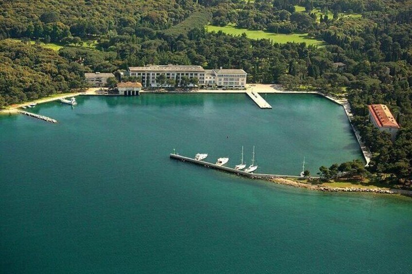 Vista aérea de un puerto deportivo con barcos amarrados, un edificio grande y un muelle que se extiende hacia una masa de agua tranquila rodeada de vegetación. 3