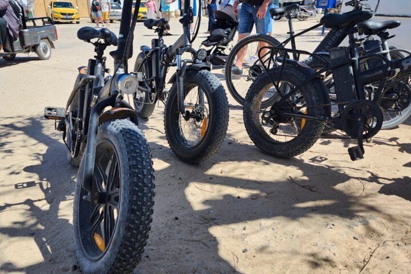 A row of electric bikes parked on sandy ground with people in the background. 4