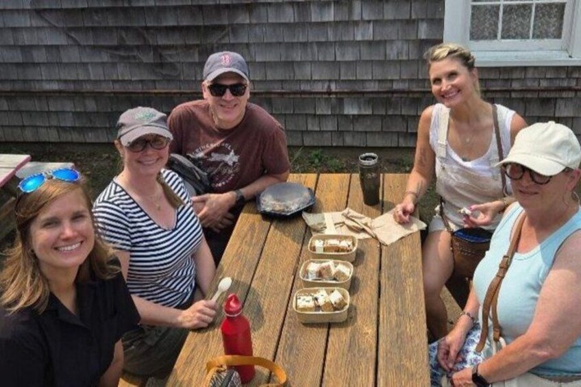 A group of people sitting at a wooden table outdoors with food and drinks. 4