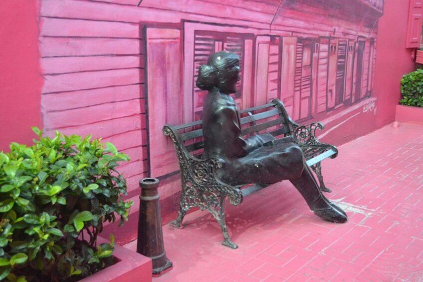 A statue of a seated figure on a bench, a pink wall with a mural, and green foliage. 2