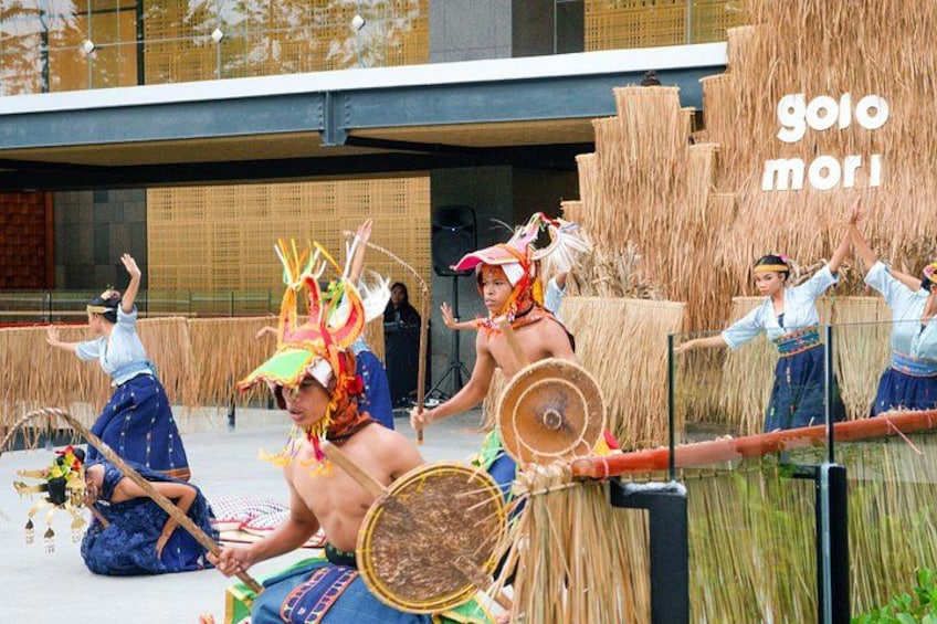 A cultural performance with dancers in traditional costumes and headgear, performing in an outdoor setting with a modern building in the background. 2