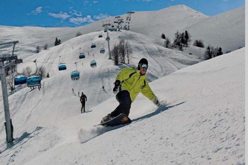 En person på snowboard styrter ned ad en sneklædt skråning med ski elevatorer i baggrunden. 2