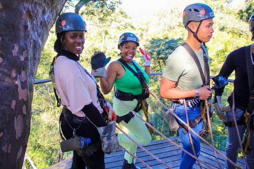 People on a zipline course, wearing helmets and harnesses, with trees and a wooden platform in the background. 3