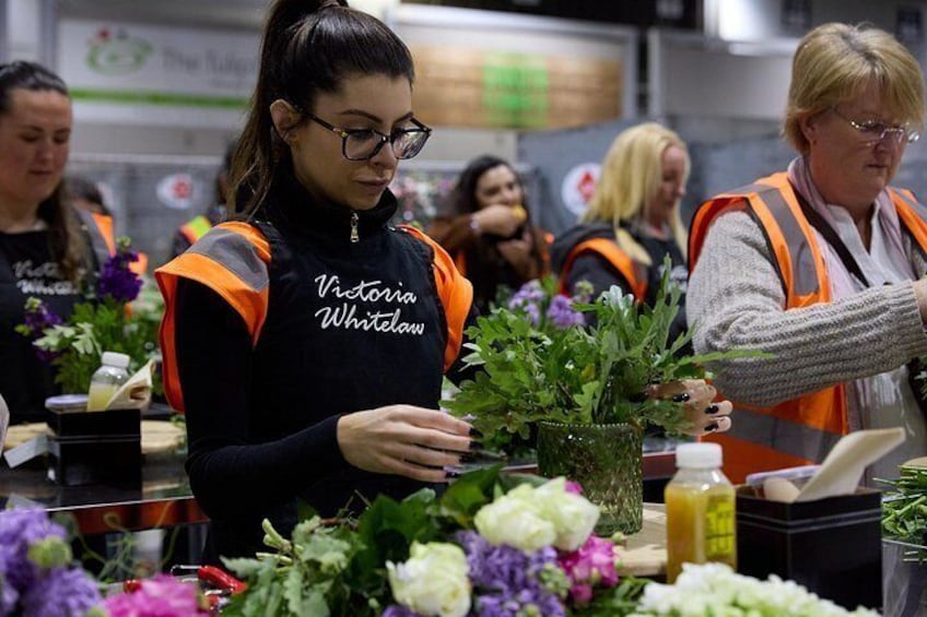 A person arranging flowers in a professional setting, likely a florist shop, with other individuals in the background. 1