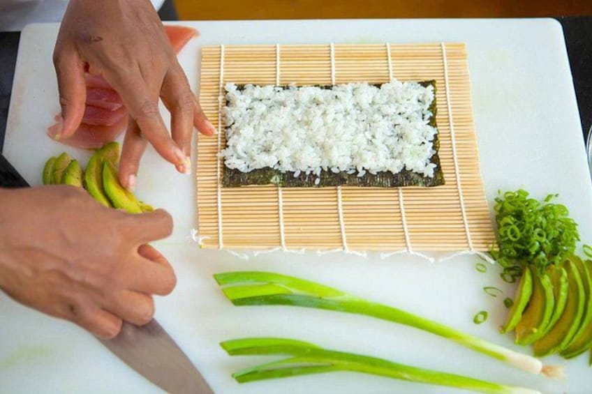 A person preparing sushi on a cutting board with rice, nori, avocado, and green onions. 3