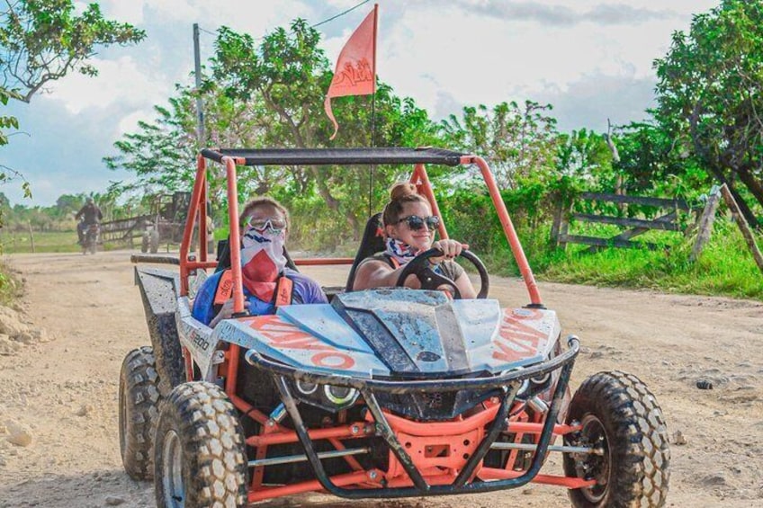 A four-wheeled vehicle with a raised platform and a flag on top, driven by two people on a dirt road with greenery in the background. 3