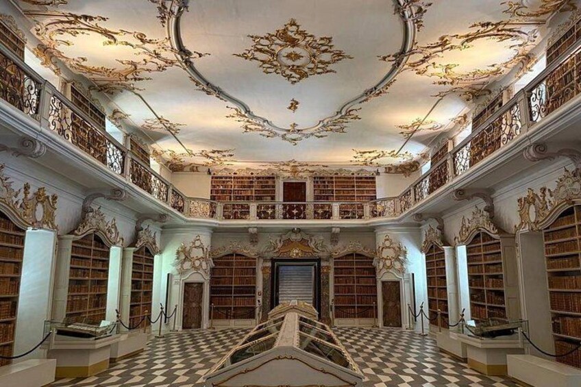 An ornate library interior with a checkered floor, elaborate ceiling frescoes, and tall bookshelves. 2