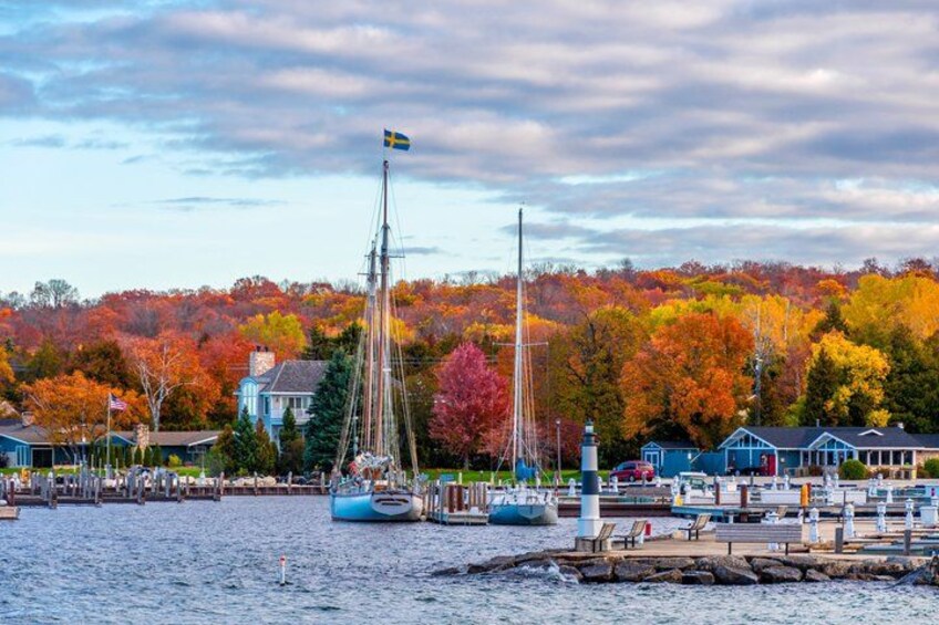 A waterfront with sailboats, a flagpole with a flag, and autumn-colored trees. 1