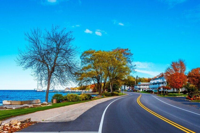 A coastal road with a clear blue sky, autumn-colored trees, and a body of water. 3