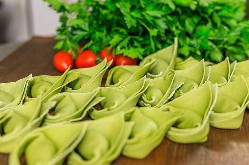 A collection of green pasta shapes on a wooden surface, with a bunch of parsley and cherry tomatoes in the background. 1
