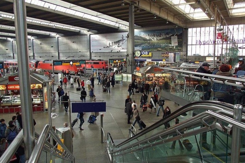 A busy indoor train station with escalators, shops, and a large window displaying a mountain landscape. 2