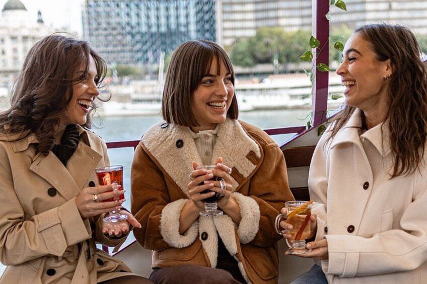 Three people sitting on a bench, holding drinks, with a cityscape in the background. 1