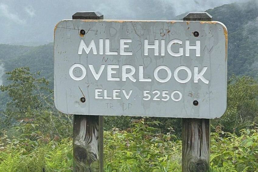 A weathered signpost with the text 'MILE HIGH OVERLOOK ELEV 5250' against a backdrop of lush greenery and mountains. 2