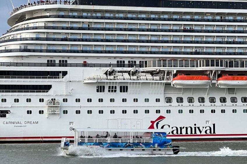Et stort krydstogtskib, Carnival Dream, med flere dæk og passagerer synlige på øverste niveauer. En mindre båd ses i forgrunden, mens den sejler gennem vandet. 1