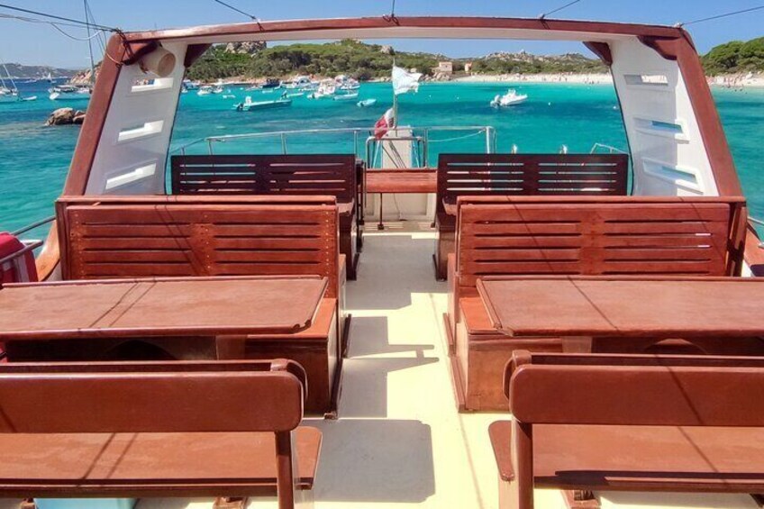 A boat deck with wooden benches and tables, a railing, and a view of turquoise waters and boats in the distance. 5