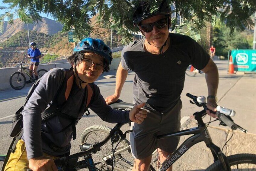 Two people on bicycles, one wearing a helmet and sunglasses, and the other holding a bike. 1